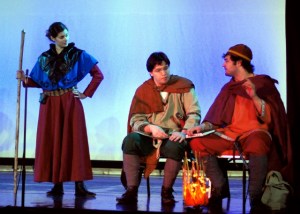 Scene from a play: two men dressed in different styles seated at a fire in conversation, while a woman holding a staff stands to the left and looks on.